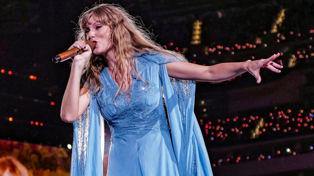 Taylor Swift with one arm outstretched and holding a microphone during a concert