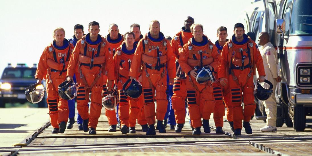 A group of astronauts in orange space suits walk across an airfield