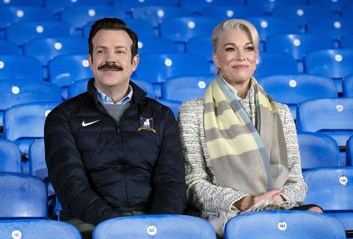 Jason Sudeikis as Ted Lasso and Hannah Waddingham as Rebecca in Ted Lasso, sitting on chairs in a football stadium